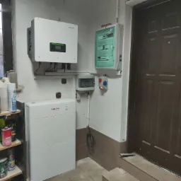 Instalacja z białym inwerterem i akumulatorem Sofar Solar w pomieszczeniu gospodarczym z półkami pełnymi pojemników i puszek, obok brązowych drzwi.