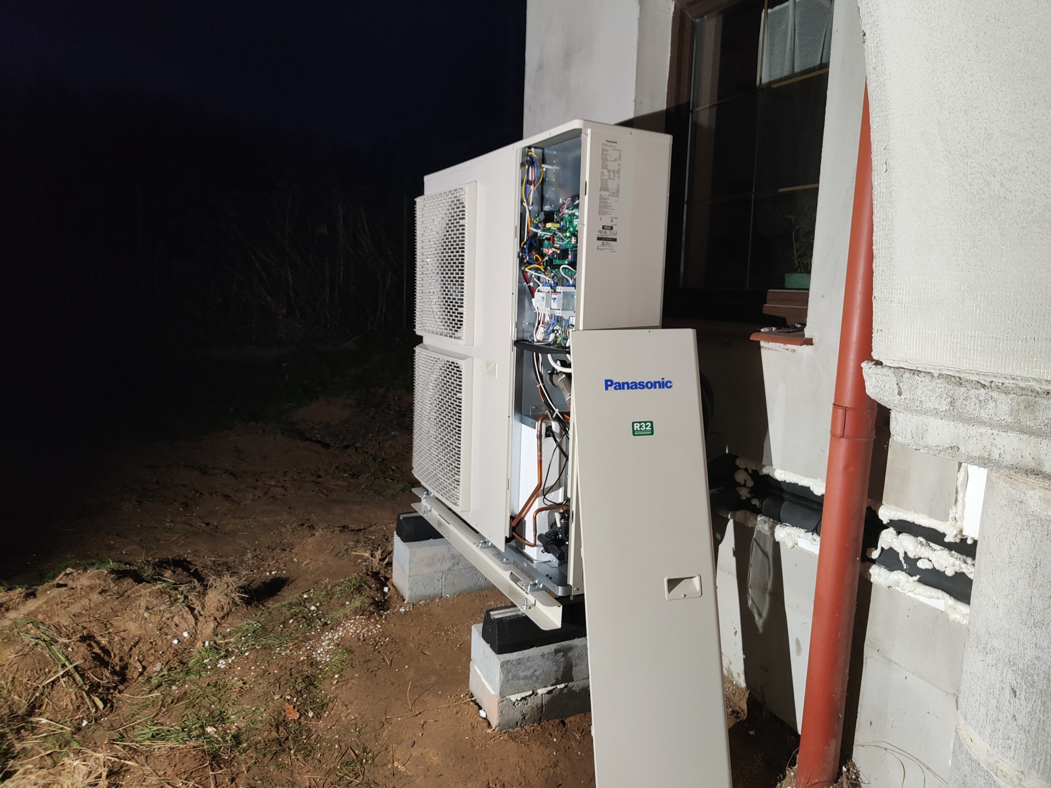 Rozłożona na części zewnętrzne jednostki pompy ciepła Panasonic R32 na betonowych blokach, z widocznymi przewodami i elektroniką, obok ściany budynku z pomarańczową rurą spustową.