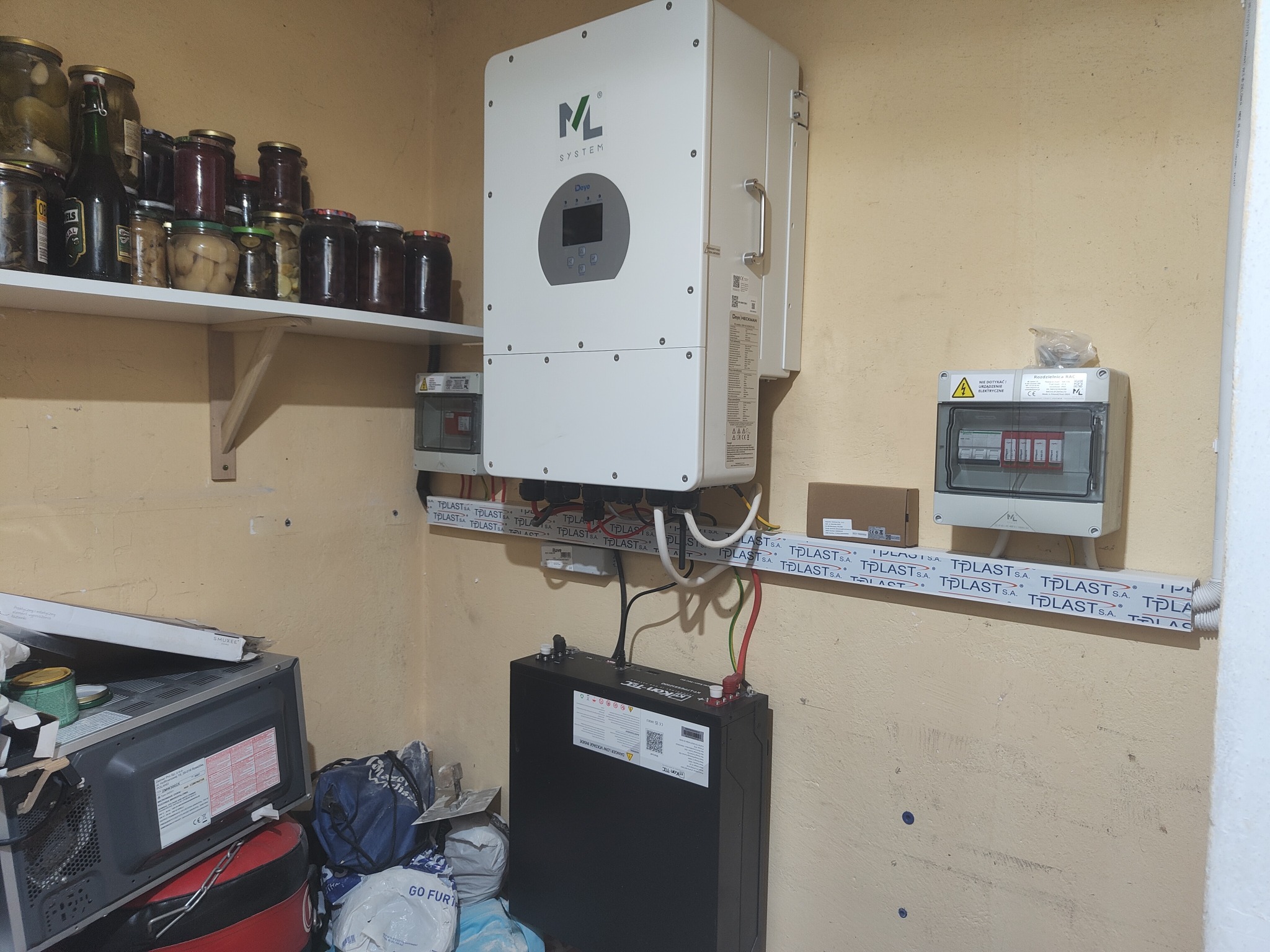 Domowa instalacja fotowoltaiczna w trakcie montażu: biały inwerter ML System na ścianie, akumulator energii na podłodze, puszki elektryczne i kable w listwach TPLAST, słoiki z przetworami na półce...