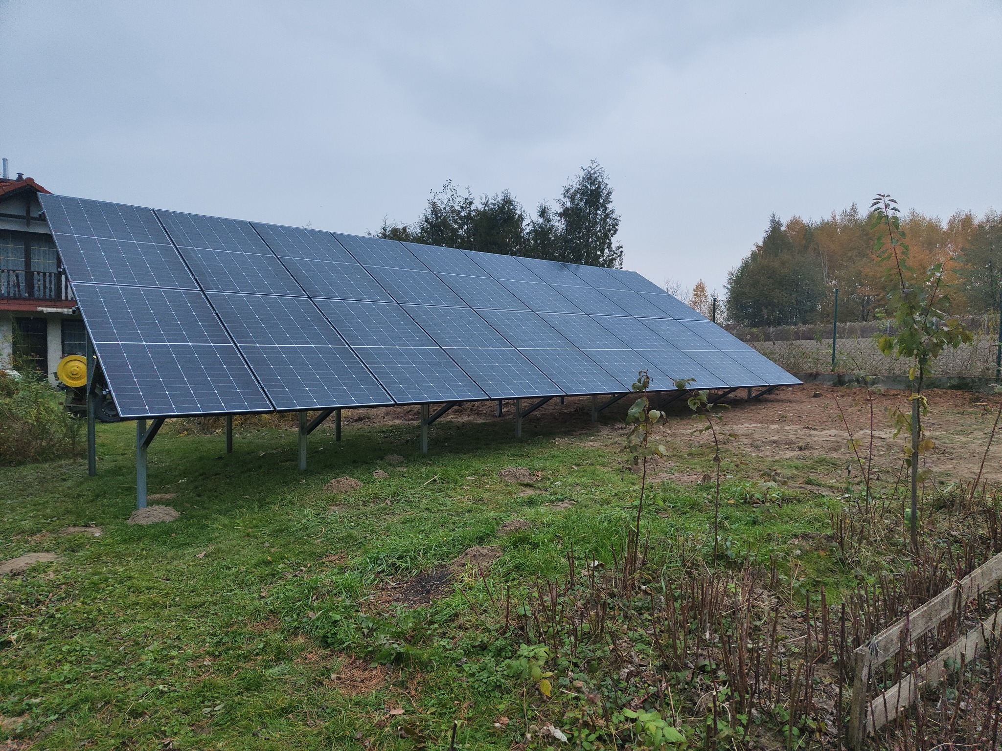 Panel słoneczny na metalowej konstrukcji, zainstalowany na trawiastym terenie obok domu, w tle drzewa i pochmurne niebo.