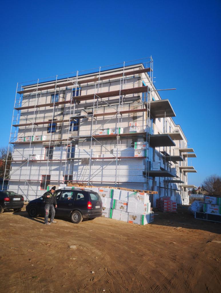 Wielopiętrowy budynek w trakcie prac, pokryty rusztowaniami i izolacją, z zapasem materiałów na placu budowy obok zaparkowanego samochodu osobowego.