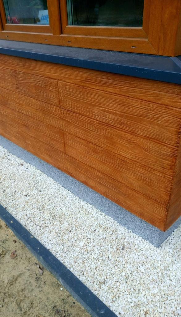 Fragment ocieplonej elewacji imitującej drewno, z widocznym oknem i obsypanym kamykami cokołem.