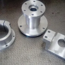 Trzy precyzyjnie obrobione, aluminiowe elementy z otworami na śruby, leżące na szarej, metalowej powierzchni z widocznymi śladami obróbki.