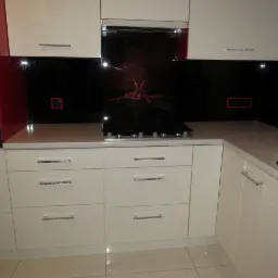 Biała zabudowa kuchenna z czarnym panelem szklanym nad blatem i płytą grzewczą, akcenty czerwieni na ścianie, minimalistyczne uchwyty szuflad.