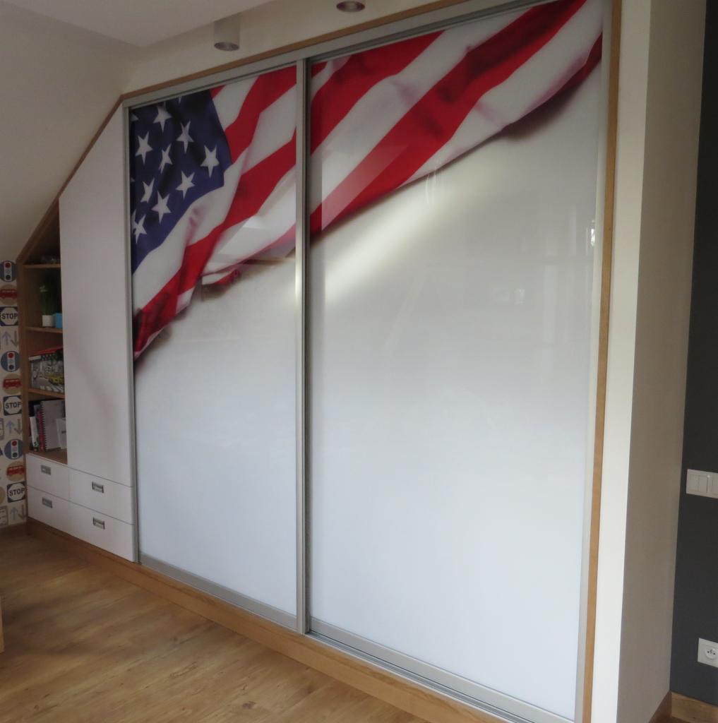 Biała szafa wnękowa z przesuwnymi drzwiami, na których nadrukowano fragment flagi USA, widoczna z boku zabudowa z półkami i szufladami, drewniana podłoga.