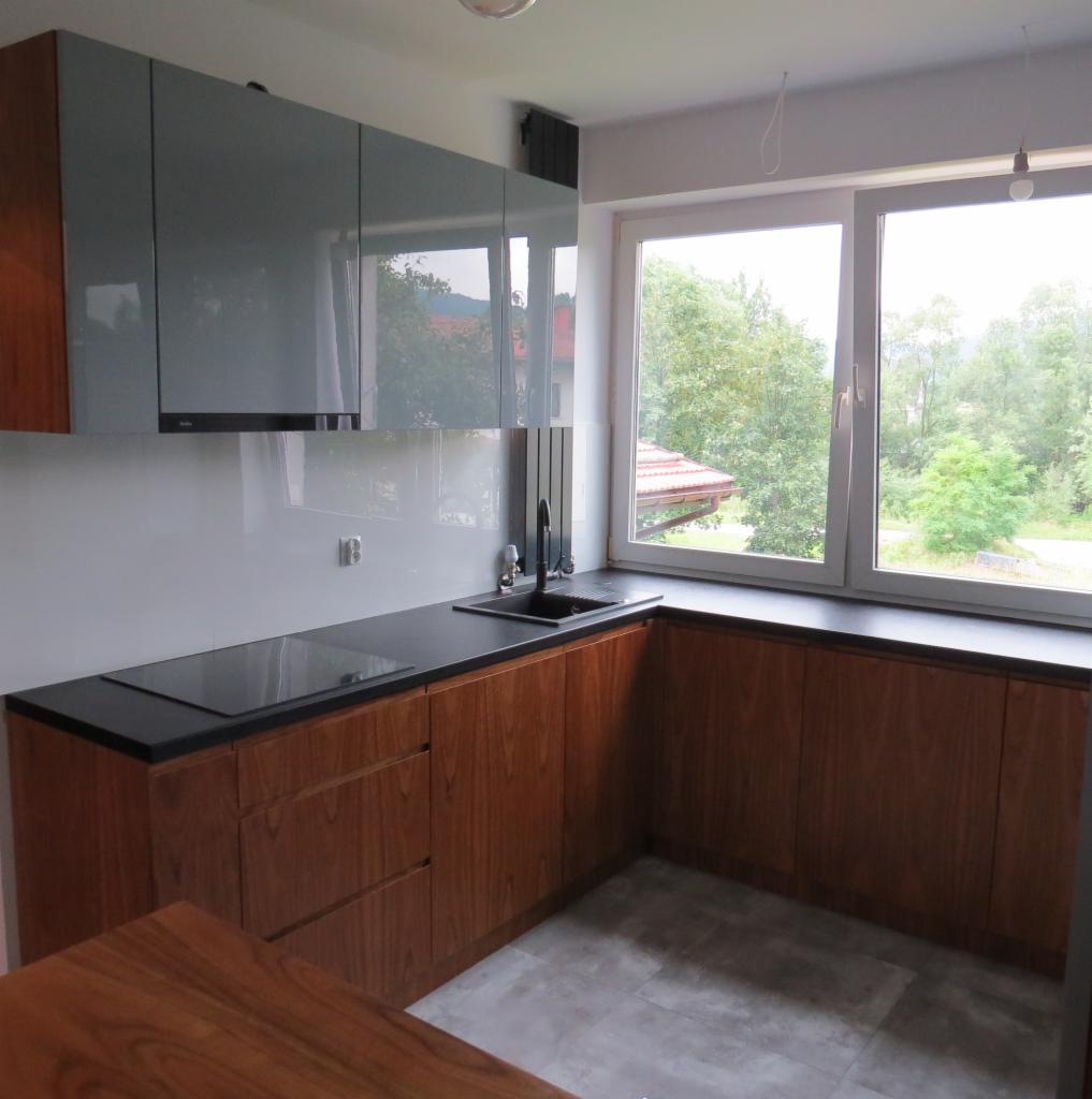 Nowoczesna kuchnia z drewnianymi szafkami, czarnym blatem i szarymi górnymi szafkami, widok na zieleń za oknem.