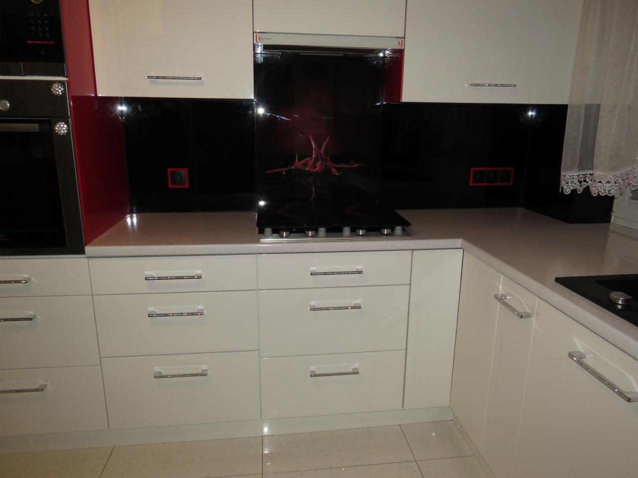 Biała zabudowa kuchenna z czarnym panelem szklanym nad blatem i płytą grzewczą, akcenty czerwieni na ścianie, minimalistyczne uchwyty szuflad.