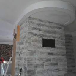 Ściana wykończona szarą płytą imitującą kamień z wnęką na telewizor, połączona z sufitem zaokrąglonym elementem gipsowym, widoczne narzędzia i materiały budowlane w tle.