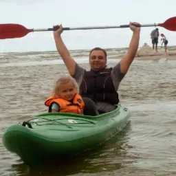 Ojciec z córką w zielonym kajaku na wzburzonym morzu, oboje w kamizelkach ratunkowych. Mężczyzna trzyma wiosło nad głową. W tle osoby budujące zamek z piasku.