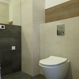 M4 Wykończenia Wnętrz Mariusz Śliwka - Nowoczesna toaleta podwieszana z chromowanym przyciskiem spłukującym, obok fragmentu ściany wykończonej płytkami imitującymi drewno, widoczne szare płytki podłogowe i fragment ściany z ciemnymi...