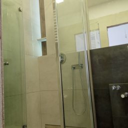 M4 Wykończenia Wnętrz Mariusz Śliwka - Nowoczesna kabina prysznicowa z deszczownicą i szklanymi drzwiami, wykończona beżowymi płytkami i ciemnoszarymi elementami, z widoczną półką w ścianie.