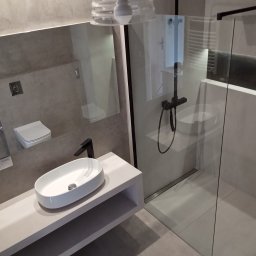 M4 Wykończenia Wnętrz Mariusz Śliwka - Nowoczesna łazienka w Tychach z owalną umywalką nablatową, czarną armaturą i szklaną kabiną prysznicową typu walk-in, oświetlona lampą z żarówką Edison.