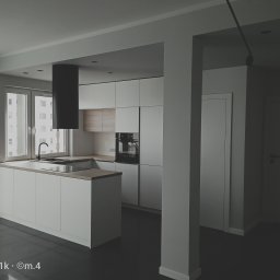 M4 Wykończenia Wnętrz Mariusz Śliwka - Nowoczesna, minimalistyczna kuchnia w odcieniach bieli i szarości z czarnym okapem na tle okna z widokiem na budynek mieszkalny.