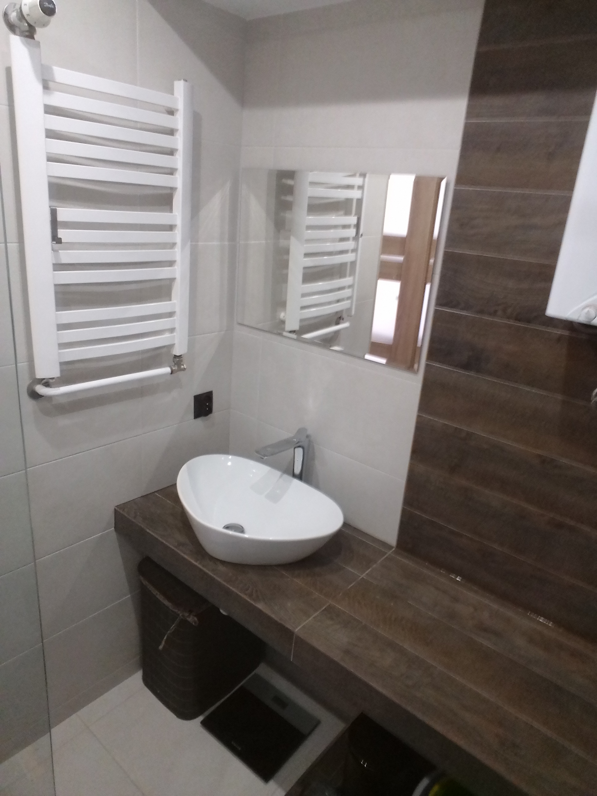 Nowoczesna łazienka z owalną umywalką nablatową, baterią chromowaną, grzejnikiem drabinkowym i brązowymi płytkami imitującymi drewno na ścianie.