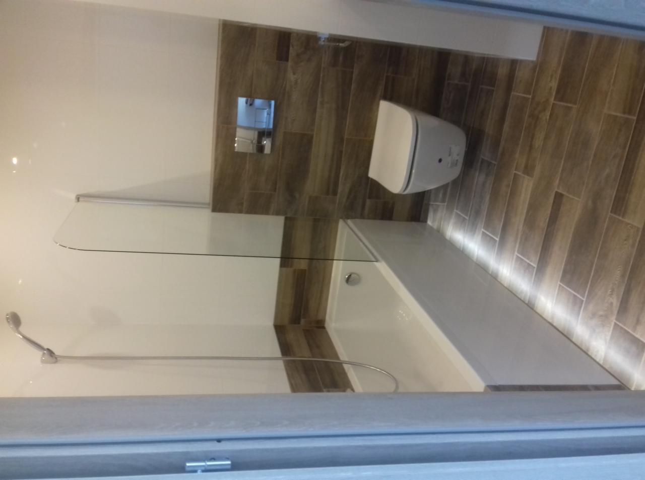 Nowoczesna łazienka z wanną, szklanym parawanem nawannowym, podwieszaną toaletą i płytkami imitującymi drewno na ścianie i podłodze, oświetlenie LED pod wanną.