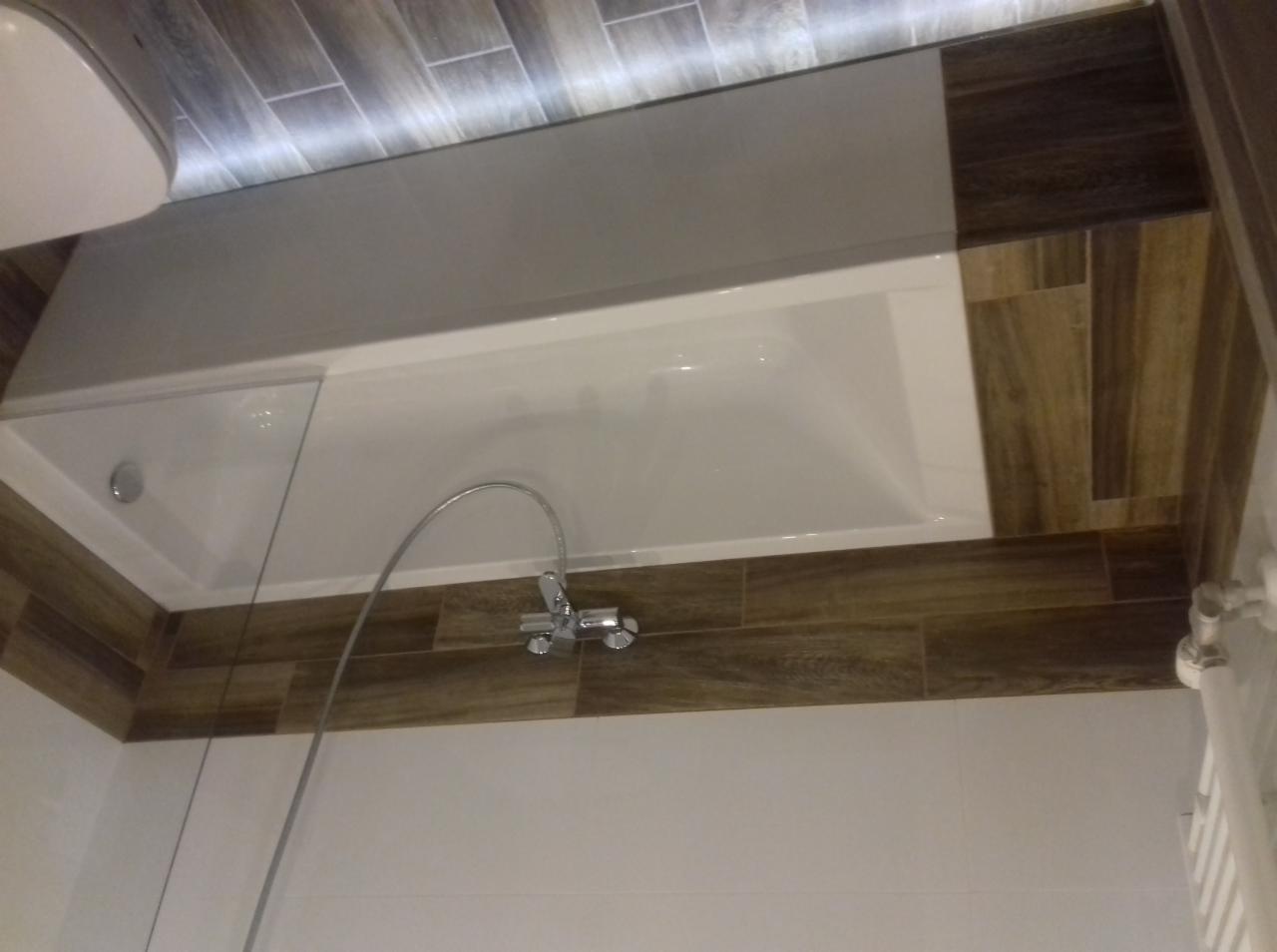 Wykończona łazienka z wanną obudowaną brązowymi płytkami imitującymi drewno, chromowana bateria wannowa, białe ściany i sufit z oświetleniem LED.