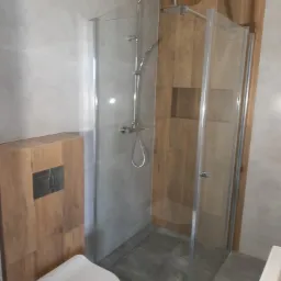 Kabina prysznicowa z deszczownicą i baterią termostatyczną, obok toaleta podwieszana z drewnianą obudową stelaża, szare płytki podłogowe i ścienne, drewnopodobna ściana wewnątrz kabiny.