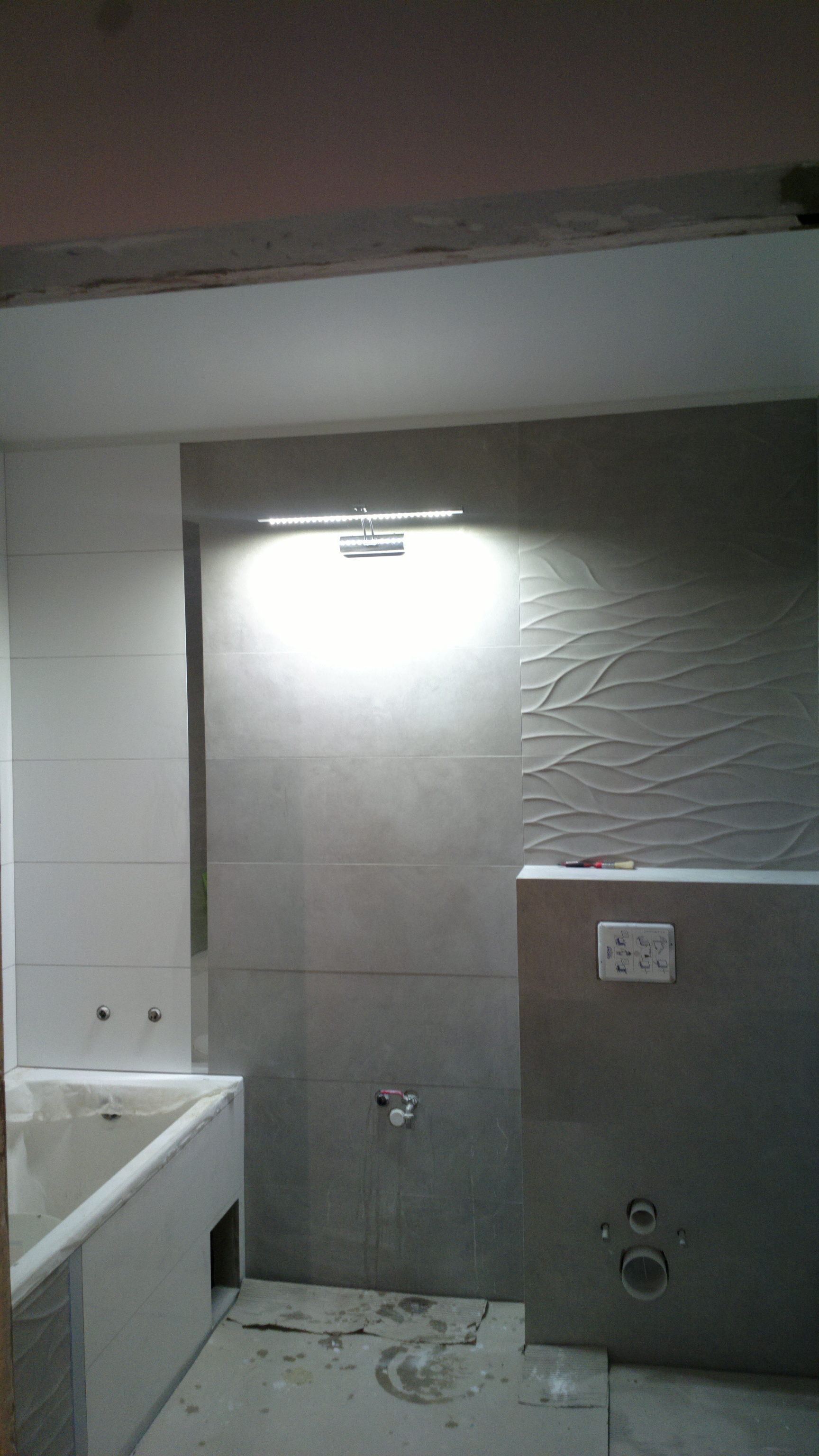 Remontowana łazienka z szarymi i białymi płytkami, widoczna wanna w zabudowie, oświetlenie LED nad umywalką i fragment podłogi z zabrudzeniami po remoncie.