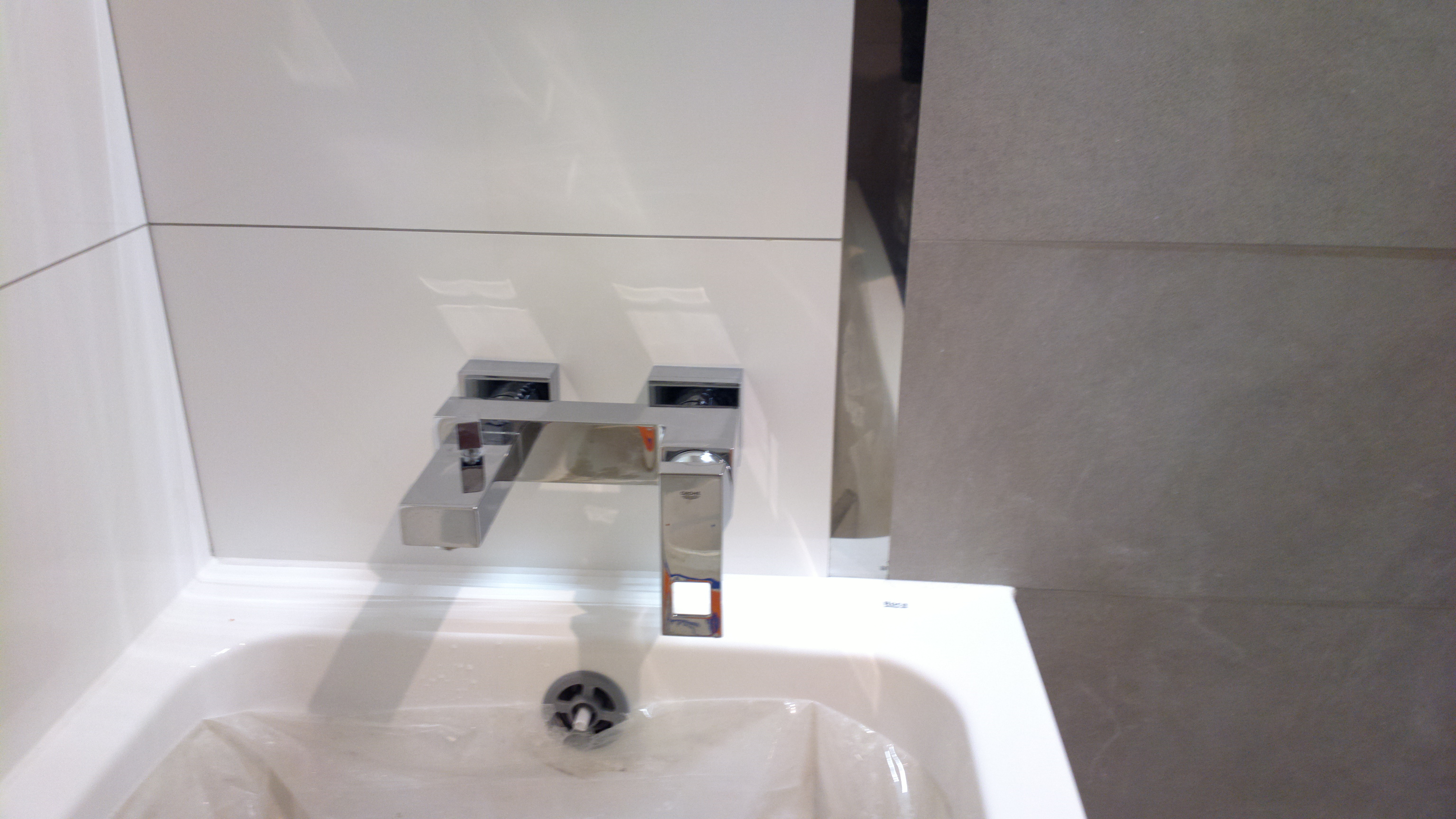 Bateria umywalkowa ścienna Grohe zamontowana nad białą umywalką, zabezpieczoną folią ochronną, na tle białych paneli ściennych i szarych płytek.