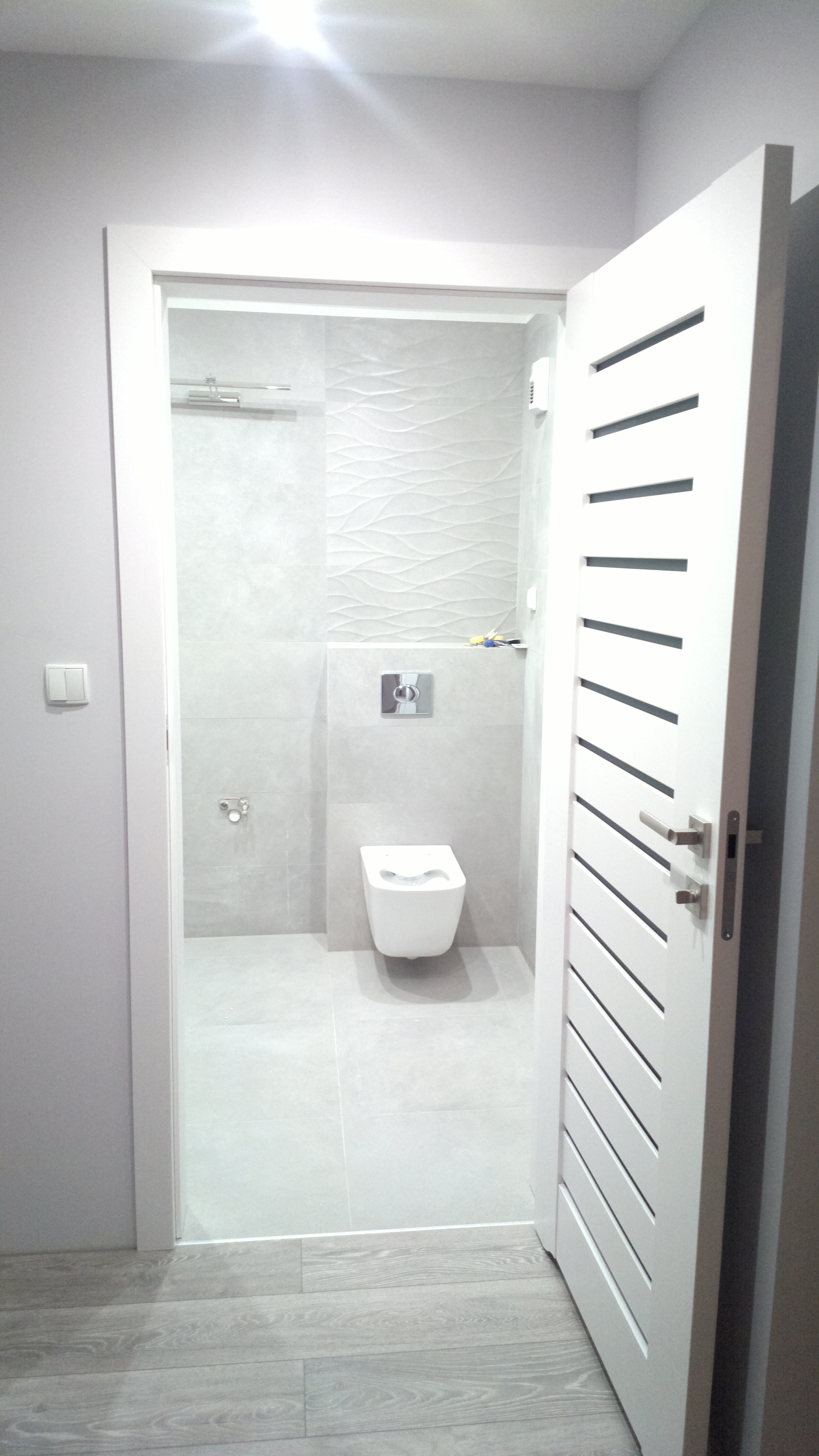 Nowoczesna łazienka w odcieniach szarości i bieli z podwieszanym bidetem, płytkami z delikatnym wzorem fal i białą, ażurową, uchyloną  drzwi.