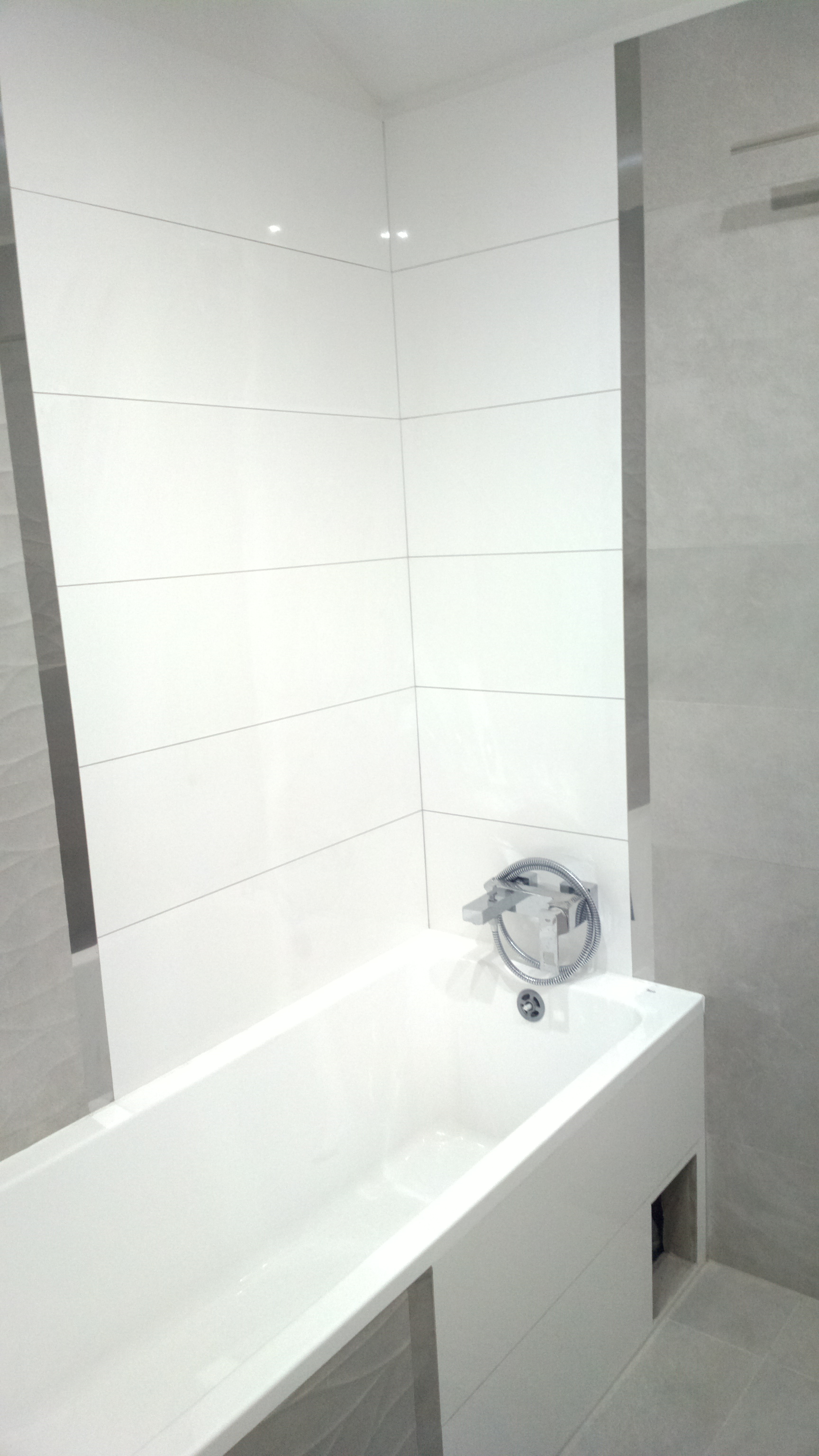 Nowoczesna biała wanna z chromowaną armaturą i szarymi płytkami na ścianach, minimalistyczny design.