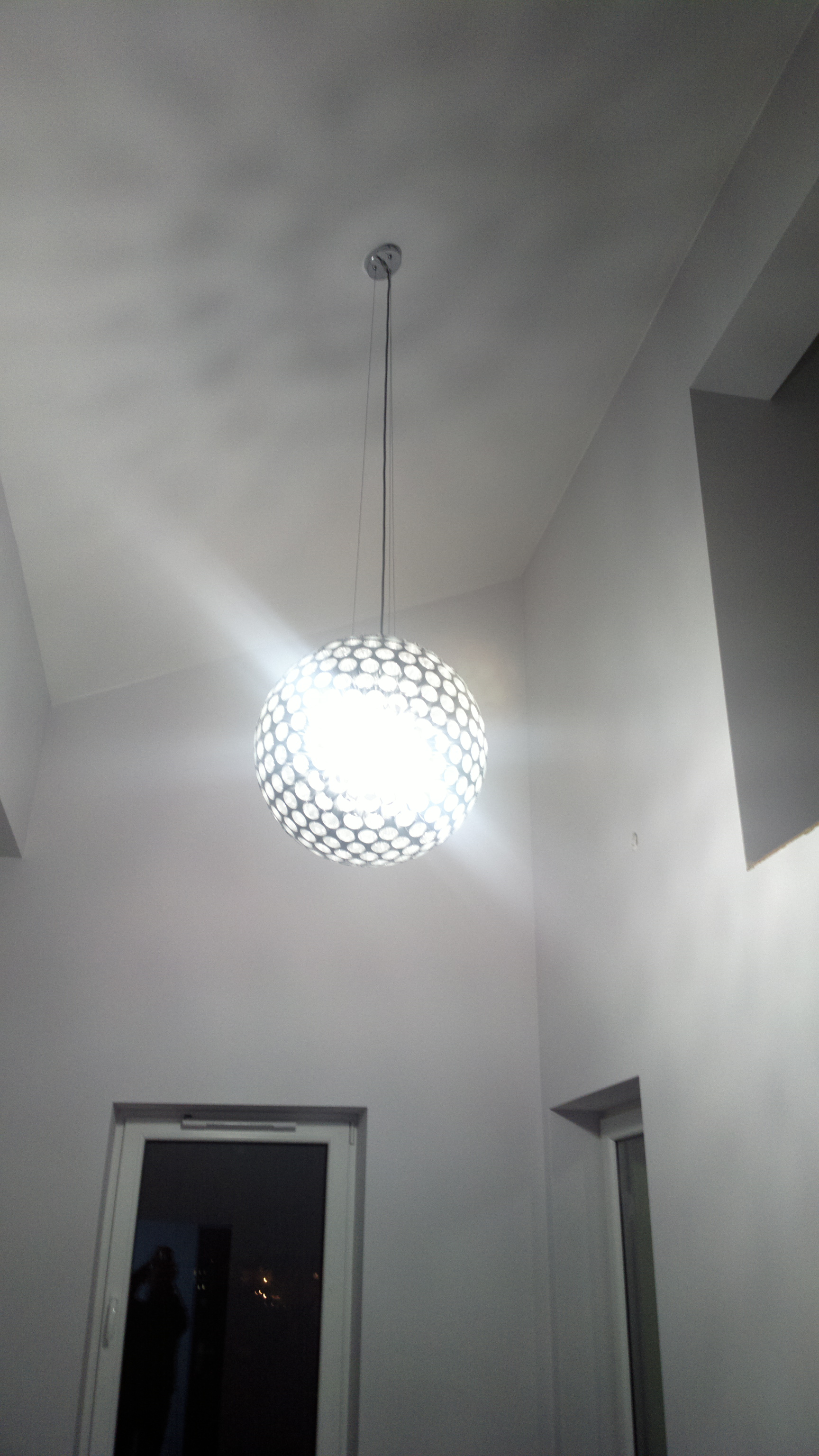 Klosz lampy wiszącej w kształcie ażurowej kuli z widoczną żarówką, na tle szarego sufitu i ścian w nowym mieszkaniu.