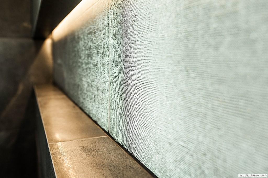 Ściana wyłożona dekoracyjną, strukturalną tapetą w odcieniach szarości, podświetlona od góry listwą LED, z niskim, ceramicznym cokolikiem w kolorze miedzi.