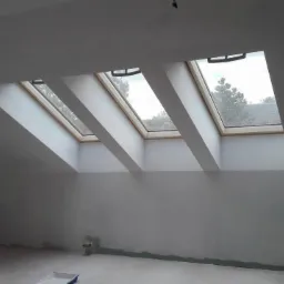 Poddasze z trzema oknami dachowymi, ściany w trakcie szpachlowania, widoczne ślady po gładzi i niedomalowane fragmenty przy podłodze, surowy stan wykończenia.