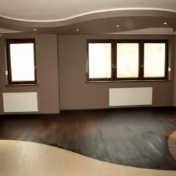 Wykończone wnętrze pokoju z ciemną drewnianą podłogą przechodzącą w jasne płytki, brązowe ściany, białe grzejniki i podwieszany sufit z oświetleniem punktowym.