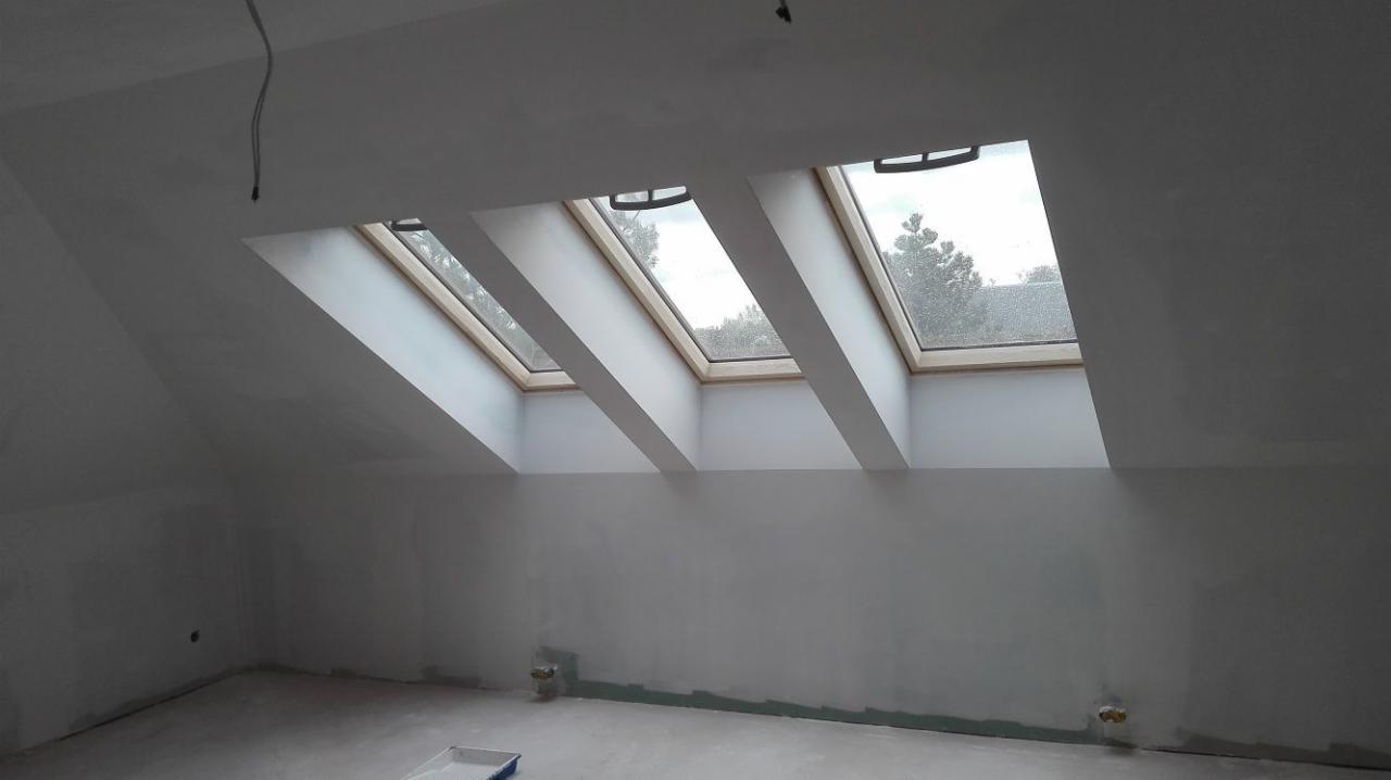 Poddasze z trzema oknami dachowymi, ściany w trakcie szpachlowania, widoczne ślady po gładzi i niedomalowane fragmenty przy podłodze, surowy stan wykończenia.