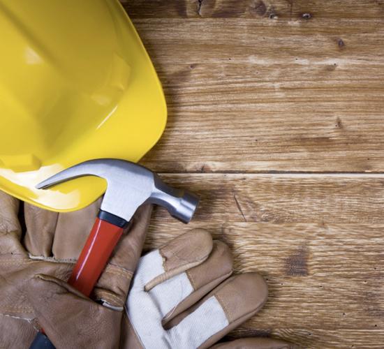 Żółty kask budowlany, młotek z czerwonym trzonkiem i skórzane rękawice robocze leżące na drewnianym blacie.