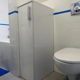 Biała szafka łazienkowa z długim, pionowym uchwytem, dopasowana do płytek z niebieskim akcentem, obok toaleta podwieszana.