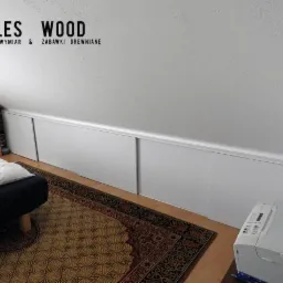 Biała zabudowa wnęki pod skosem ze przesuwnymi drzwiami, widok z boku ł&oacute;żka i dywanu w pokoju na poddaszu, logo firmy widoczne na ścianie.