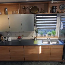 Jasna kuchnia z drewnianymi frontami szafek dolnych i szarymi górnymi, z oknem w tle, na parapecie którego stoją doniczki z ziołami. Na blacie widoczne owoce, blender i drobne sprzęty AGD.