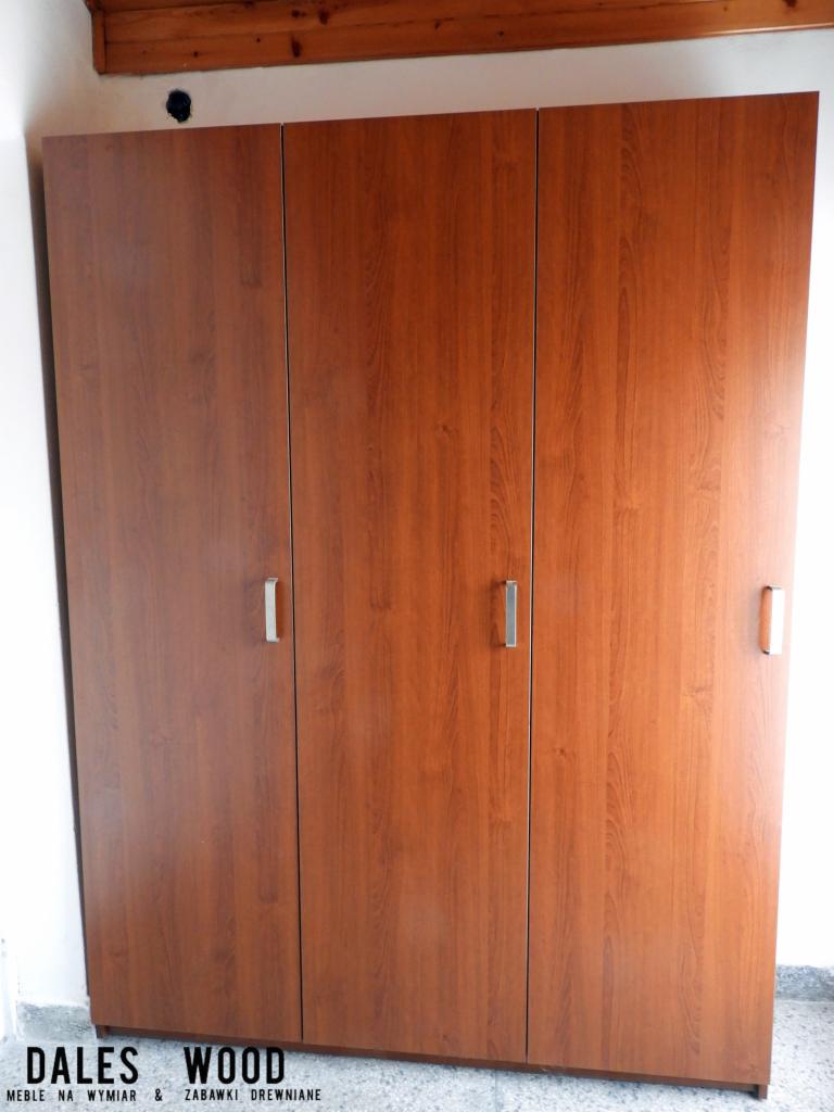 Trzydrzwiowa szafa z drewnopodobnej płyty z podłużnymi, metalowymi uchwytami, widoczna od frontu, z fragmentem drewnianego sufitu w tle.