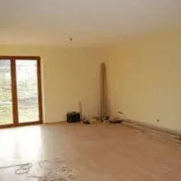 Pusty pokój w trakcie przygotowań do prac wykończeniowych, widoczne ściany pokryte jasnym podkładem, drewniana podłoga, drabina oparta o ścianę, przewody elektryczne na podłodze oraz lampa...