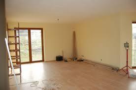 Pusty pokój w trakcie przygotowań do prac wykończeniowych, widoczne ściany pokryte jasnym podkładem, drewniana podłoga, drabina oparta o ścianę, przewody elektryczne na podłodze oraz lampa...