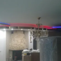 Salon w trakcie prac wykończeniowych w Białej Podlaskiej, widoczny kamienny kominek, ozdobny żyrandol oraz podświetlany sufit z elementami mozaiki i listwami LED w kolorach czerwonym i niebieskim.