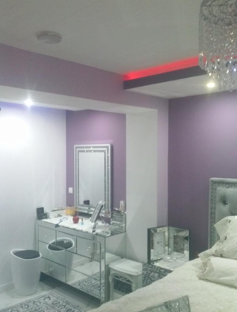 Liliowa sypialnia z lustrzaną komodą, kryształowym żyrandolem i czerwonym oświetleniem sufitowym.