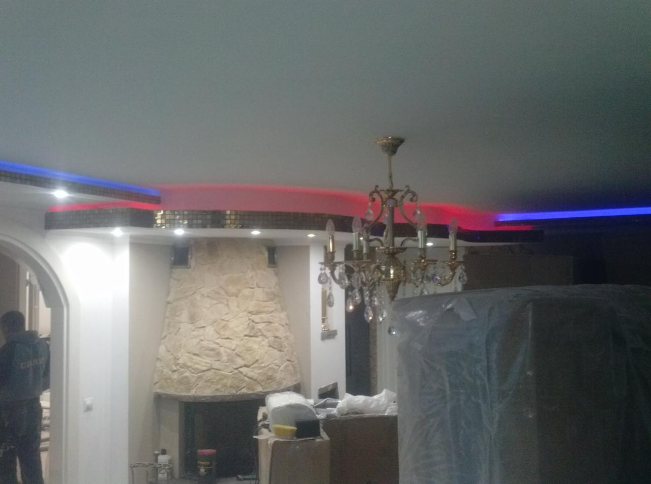 Salon w trakcie prac wykończeniowych w Białej Podlaskiej, widoczny kamienny kominek, ozdobny żyrandol oraz podświetlany sufit z elementami mozaiki i listwami LED w kolorach czerwonym i niebieskim.