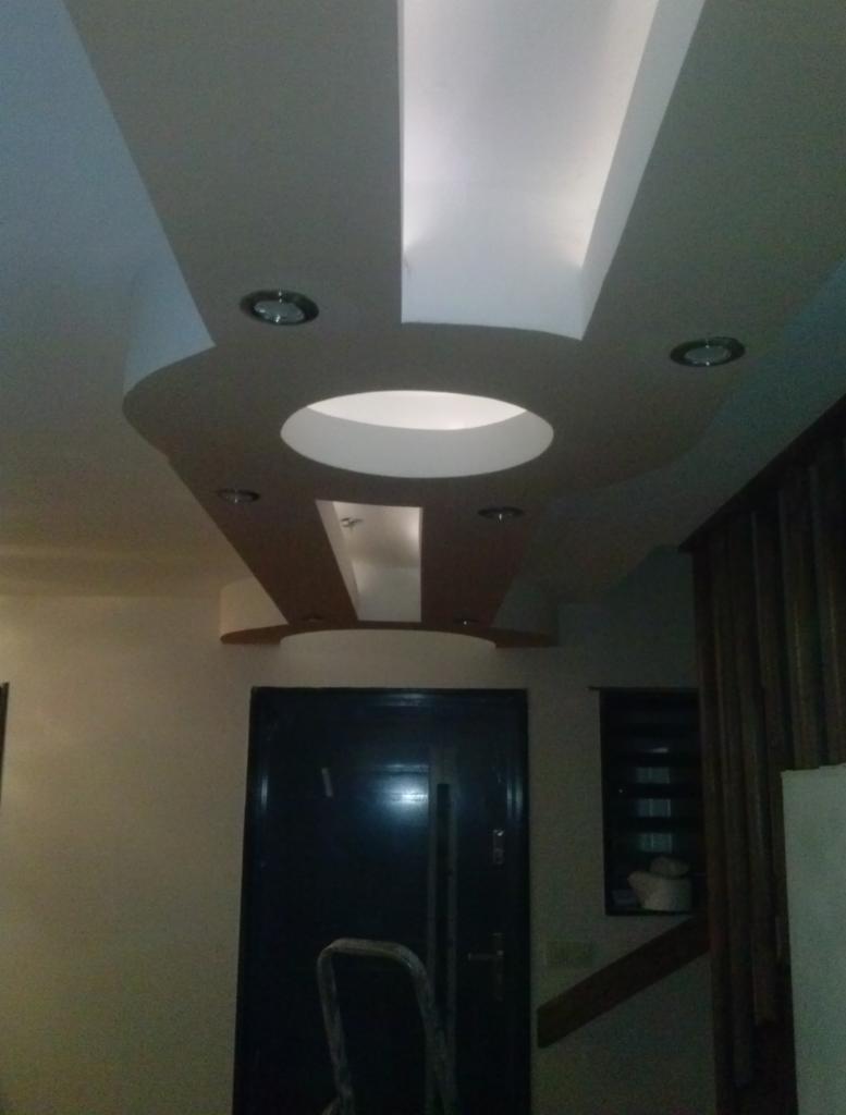 Dekoracyjny sufit podwieszany z oświetleniem punktowym i centralnym okrągłym otworem, widoczna drabina oparta o ciemne drzwi, fragment ściany i żaluzje w tle.