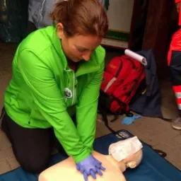 Instruktorka w zielonej bluzie demonstruje resuscytację krążeniowo-oddechową na manekinie z zabandażowaną głową, używając niebieskich rękawic ochronnych, w tle plecak i częściowo widoczny ratownik...