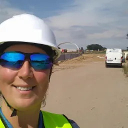 Selfie kobiety w białym kasku i odblaskowej kamizelce na terenie budowy, z odblaskami budowy w okularach przeciwsłonecznych, widoczny kontener biurowy i pojazd firmy Viacon.