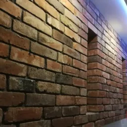 Ściana z cegły klinkierowej wewnątrz pomieszczenia z wnękami, oświetlona ledowym światłem na suficie, widoczne logo firmy w prawym dolnym rogu.