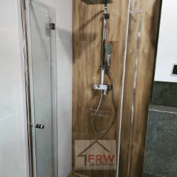 Kabina prysznicowa z chromowaną armaturą na tle płytek imitujących drewno, widoczne logo firmy remontowo-wykończeniowej w dolnym rogu.