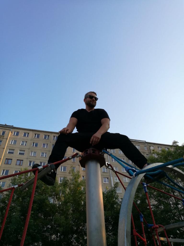 Mężczyzna w czarnym stroju i okularach przeciwsłonecznych siedzi na szczycie metalowej konstrukcji placu zabaw na tle bloku mieszkalnego i błękitnego nieba.