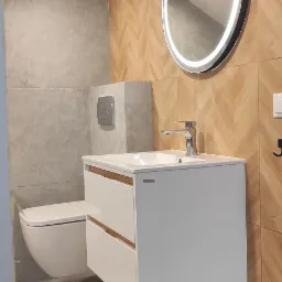 Nowoczesna łazienka z wiszącą szafką umywalkową z dwoma szufladami z drewnianymi akcentami, okrągłe podświetlane lustro na pasku, wisząca toaleta i płytki w dwóch odcieniach: drewnopodobne ułożone...