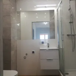 Nowoczesna łazienka z kabiną prysznicową, umywalką z szafką i toaletą, wykończona płytkami w odcieniach bieli i szarości z drewnopodobną podłogą, widoczne chromowane elementy armatury.