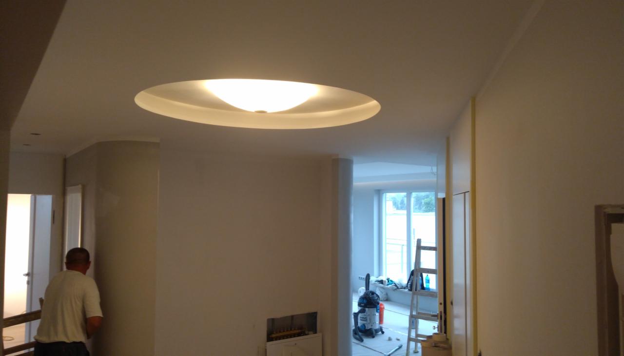 Wnętrze po malowaniu ścian z widoczną lampą sufitową w kształcie elipsy, kolumną i fragmentem pracującego glazurnika w tle.