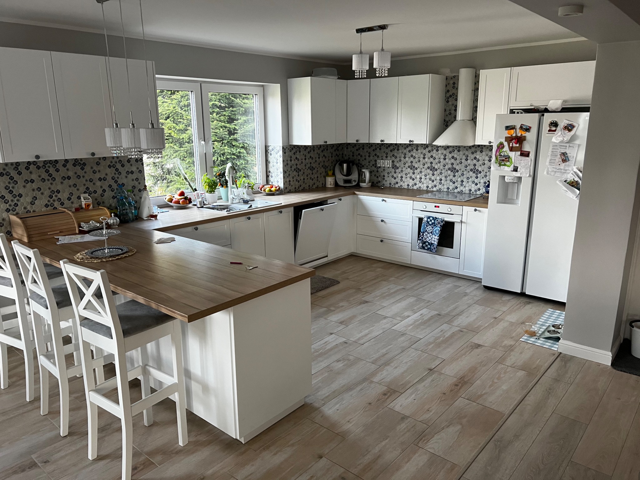 Jasna kuchnia z wyspą, białymi szafkami i drewnianym blatem, mozaika na ścianie, okno z widokiem na zieleń, drewnopodobne płytki na podłodze.